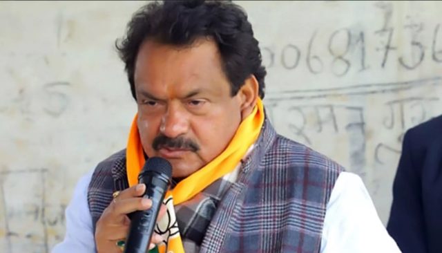 Satyapal Singh Baghel speaking into a hand-held microphone outdoors, wearing a saffron scarf and checked jacket