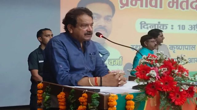 Satyapal Singh Baghel addressing a meeting from a flower-decorated dais with a Hindi banner behind him