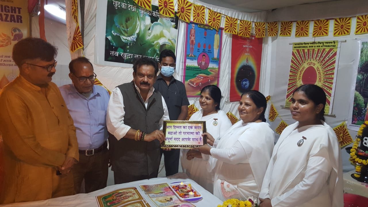 Satyapal Singh Baghel holding a framed Hindi message presented by women of the Brahma Kumaris in white, at an exhibition decorated with posters