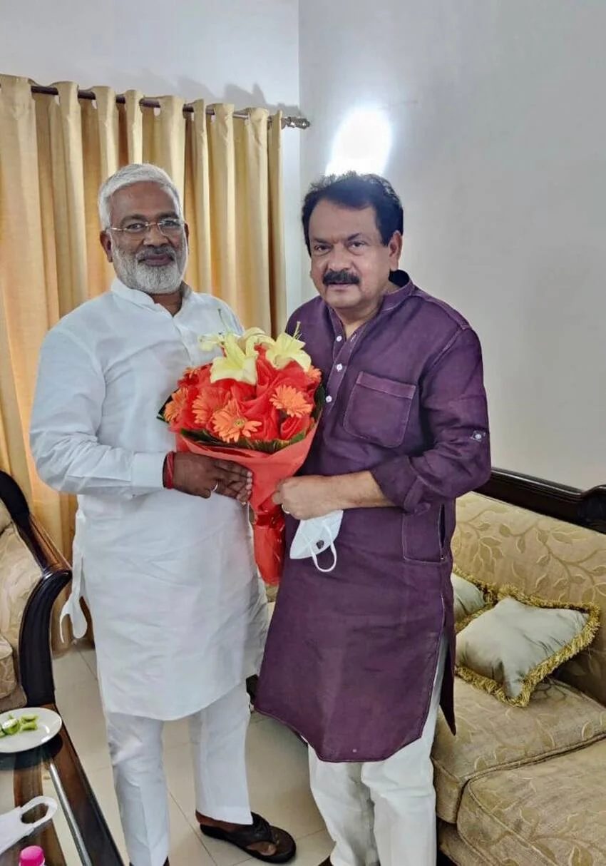Satyapal Singh Baghel, in a maroon kurta, receiving a bouquet of flowers from a man in white in a living room